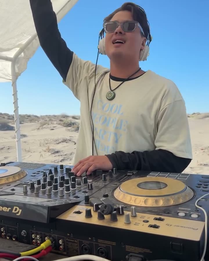 Valdi with his arm raised at a beach DJ booth in the sand