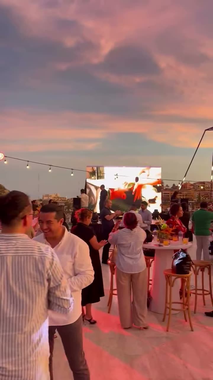A crowd at a rooftop event at sunset under string lights, with a big screen behind them