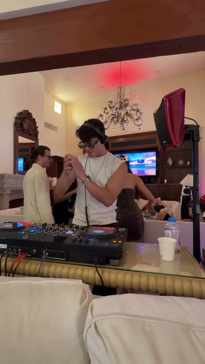 Valdi at the decks in a villa living room under colored party lights, a guest dancing beside him