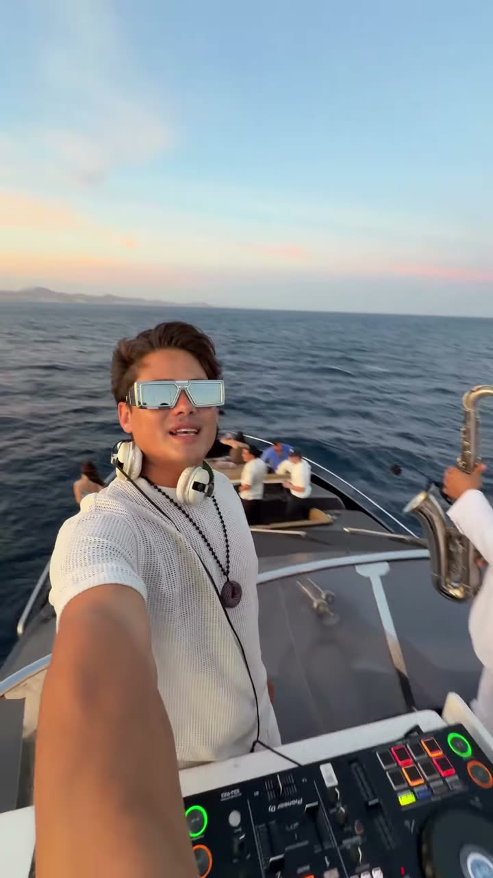 Valdi at his decks on a yacht at sea while a saxophonist in white plays beside him